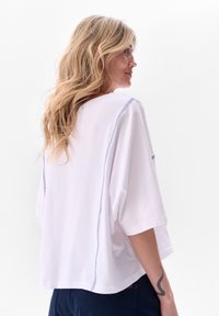 T-shirt blanc oversize à manches courtes, avec des coutures bleu contrastantes le long des coutures. Tissu en coton doux, coupe décontractée.