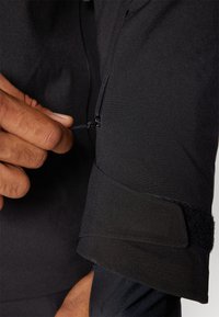 Ručně tahání malého zipu s očkem na černém látkovém rukávu s nastavitelným suchým zipem.