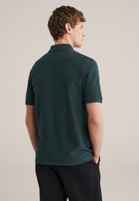 Man met korte krullende haren, gekleed in een donkergroen poloshirt met korte mouwen en een zwarte broek, staand met handen in de zakken, met de rug naar voren.
