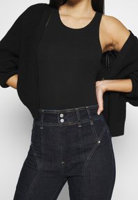 Top de tirantes negro acanalado combinado con un cárdigan negro de punto, llevado con jeans de mezclilla oscura de tiro alto que presentan costuras amarillas y botones plateados.
