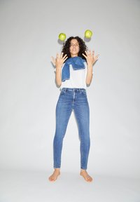 Young woman with curly hair barefoot, wearing white T-shirt and jeans, juggling two green apples against a plain gray background.