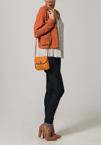 Forbrændt orange strikket cardigan over en grå mønstret top, kombineret med mørke skinny jeans og tan farvede højhælede sko. Lille orange crossbody taske.