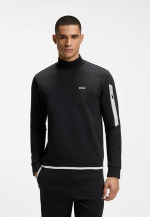 Uomo che indossa una maglia nera a maniche lunghe con logo bianco "BOSS" sul petto e dettaglio di zip bianca sulla manica sinistra, in piedi con le mani in tasca.