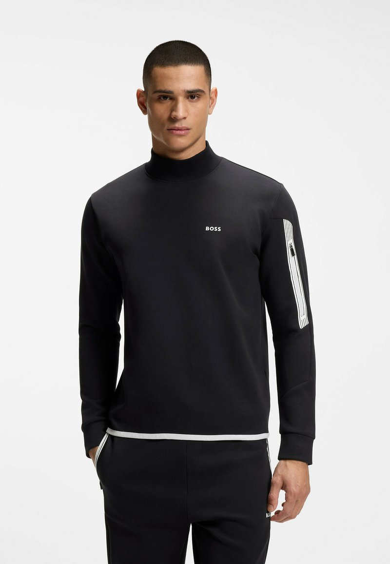 Homme portant une chemise noire à manches longues avec le logo blanc "BOSS" sur la poitrine et un détail de fermeture éclair blanche sur la manche gauche, debout les mains dans les poches.
