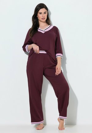 Vrouw draagt een donkerpaars tweedelig loungset met witte en roze gestreepte rand, staande op blote voeten tegen een witte muur.