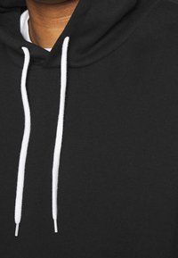 Haut à capuche noir avec une texture douce, cordons de serrage blancs avec des embouts en argent, et une capuche, mettant en avant un design décontracté.