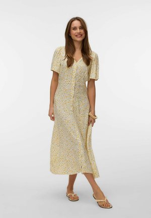 Kvinde smilende og gående i en lysegul blomsterprintet knapmidikjole med puffede ærmer, sandaler og et armbånd på hvid baggrund.