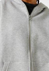 Primer plano de una sudadera con cremallera de color gris claro con cremallera plateada, llevada sobre una camiseta negra, mostrando parte del cuello y los hombros.