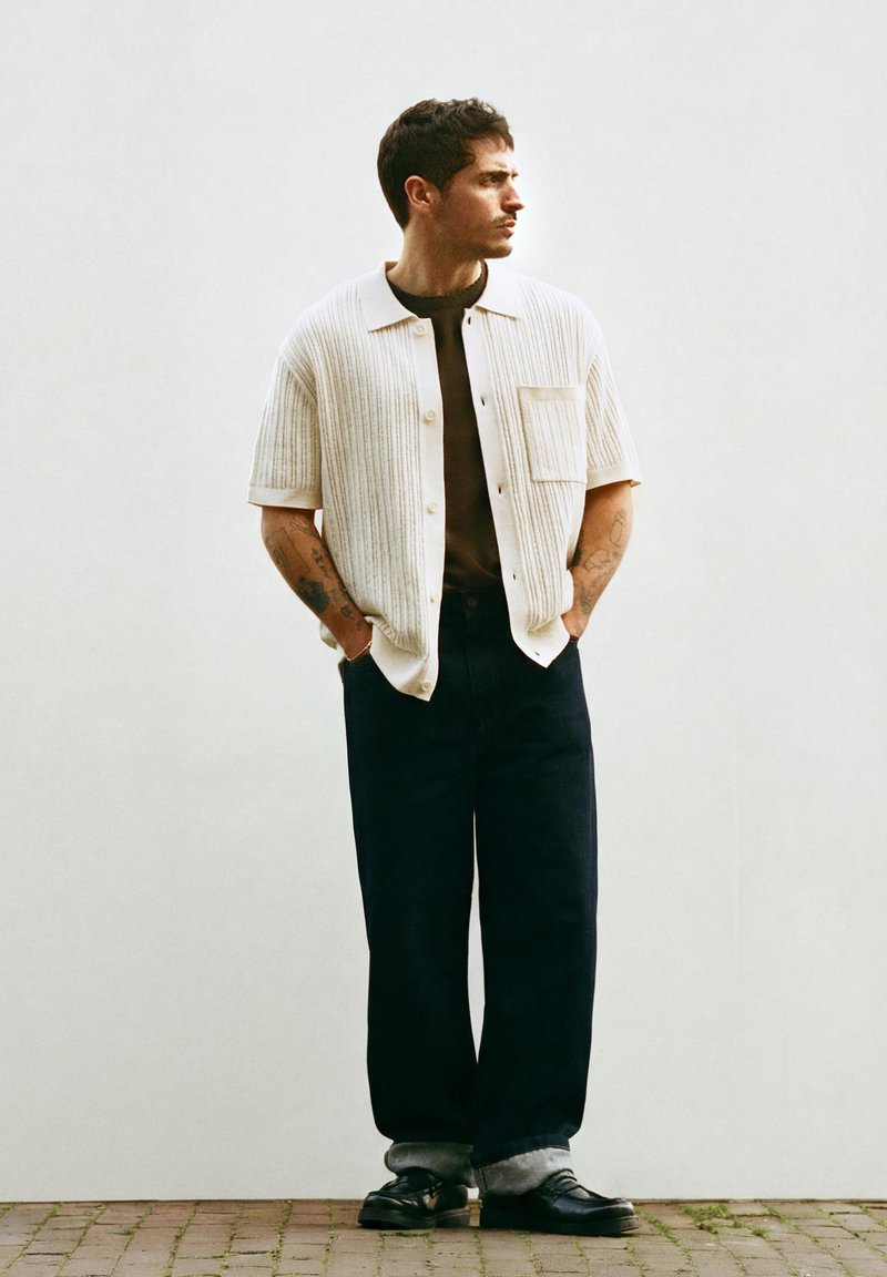 Homme debout, les mains dans les poches, portant un cardigan crème à manches courtes sur une chemise noire, un pantalon large noir, des chaussures noires et des tatouages visibles sur les bras.