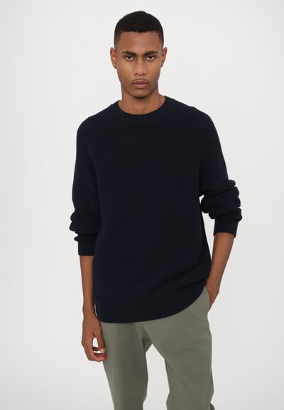 Pull en maille bleu marine avec col rond côtelé et manches longues, présentant un motif texturé et une coupe décontractée. Associé à un pantalon vert clair.