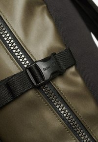 Le matériau du sac à dos vert olive présente une fermeture éclair noire, une sangle texturée et une boucle en plastique gravée de "Super".