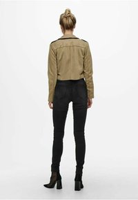 Kvinna med blont hår i en knut, iklädd en beige kort jacka, svarta slim fit jeans och svarta klackstövlar, stående med ryggen mot kameran framför en vit bakgrund.