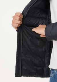 Personne portant un jean bleu et une chemise blanche tient ouverte une veste noire matelassée, montrant la poche intérieure avec une main à l'intérieur.