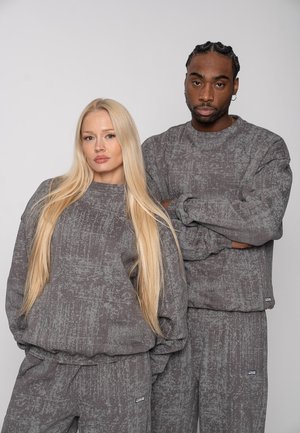 Two models, one female with long blonde hair and one male with braided hair, wearing matching gray textured sweatshirts and pants, posing against white background.