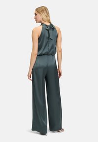 Green satin jumpsuit with wide legs, halter neck, and a tied bow at the back. Smooth texture with a relaxed fit throughout.