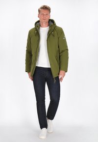 Olivgrön vadderad jacka med huva, dragkedja framtill och grå foder, i kombination med mörka jeans och vita sportskor, stående mot en vit bakgrund.