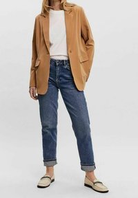 Kamelinsin takki, jossa on viistot läpät ja kaksi etutaskua, yhdistettynä vaaleansinisiin käännettyihin farkkuihin ja kermaisiin loafereihin, joissa on ketjuyksityiskohta.