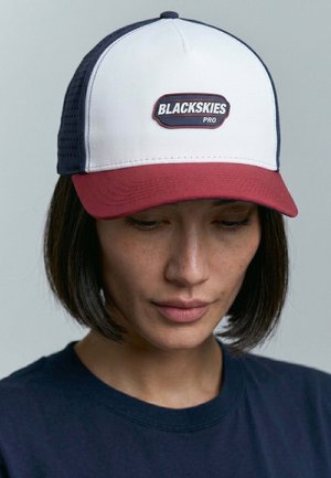 Woman with short dark hair wearing a navy shirt and a white, navy, and red cap with "BLACKSKIES PRO" logo on the front.
