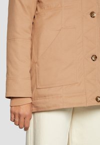 Světle hnědá prošívaná bunda s knoflíkovým zapínáním, žebrovanými manžetami a velkými kapsami. Vyznačuje se kontrastním prošíváním a hladkou, texturovanou látkou.