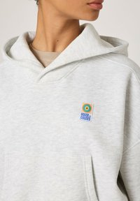 Šviesiai pilkas džemperis su gobtuvu, pagamintas iš minkštos medvilnės, su priekiu kišene, raišteliu ant gobtuvo ir spalvingu „House of Colors“ logotipo antspaudu ant krūtinės.