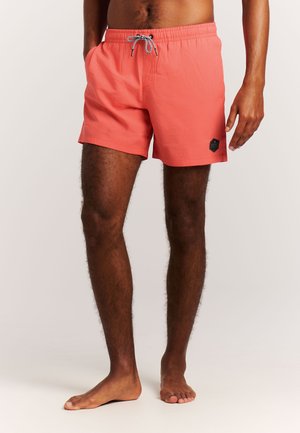 Homme debout pieds nus portant un short de bain corail avec un cordon noir et blanc, mains détendues, sur fond uni.