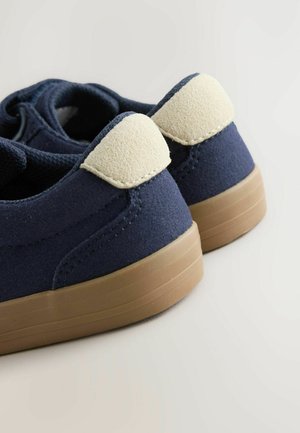 Marineblå sneakers med beige ruskind hælkapsler og brune gummisåler, vinklet for at vise bagsiden og detaljer om materialet og syningerne.