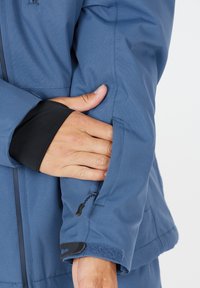 Blå jacka som visar en slät tygtextur, åtsittande ärmslut, hand i fickan och en dragkedjedetalj på ärmen. Har justerbar hårdvara.