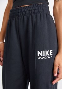 Pantaloni della tuta neri con elastico in vita, con logo 'NIKE' bianco sul lato sinistro. Il tessuto appare morbido con una texture liscia.