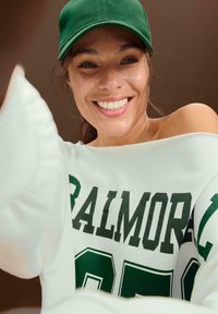 Felpa bianca oversize con testo verde "BALMORE 95" e un cappellino da baseball verde. Tessuto liscio, stile off-shoulder, vestibilità casual.