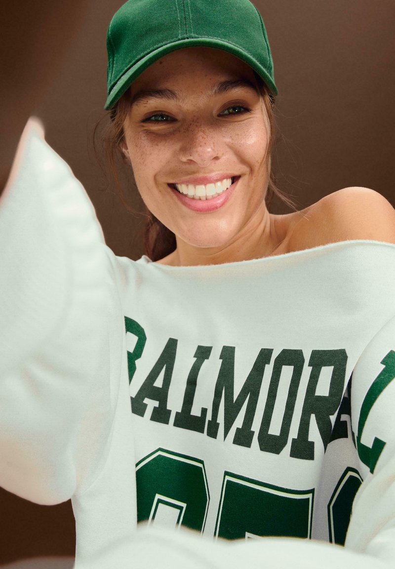 Felpa bianca oversize con testo verde "BALMORE 95" e un cappellino da baseball verde. Tessuto liscio, stile off-shoulder, vestibilità casual.