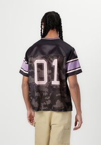 Krátký rukáv dres v černé barvě se vzorem camouflage. Obsahuje fialové detaily, číslo "01" na zadní straně a kontrastní pruhované rukávy.