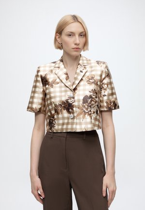 Femme portant un chemisier à manches courtes beige et marron à carreaux avec de grands imprimés floraux et un pantalon marron taille haute, debout devant un fond uni.