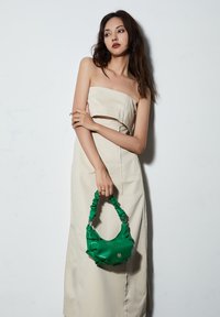 Grüner Gummihandtasche mit einem gerafften Griff; glatte Textur und abgerundete Form. Das Modell trägt ein beiges, trägerloses Kleid. Der Hintergrund ist weiß.