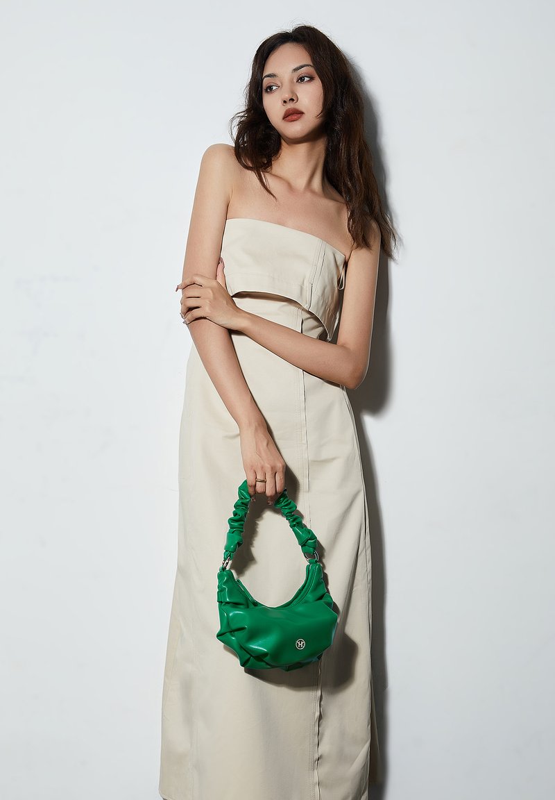 Grüner Gummihandtasche mit einem gerafften Griff; glatte Textur und abgerundete Form. Das Modell trägt ein beiges, trägerloses Kleid. Der Hintergrund ist weiß.