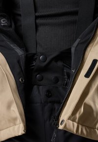 Schwarze und tanfarbene Jacke mit Reißverschluss und Druckknopfverschluss, ausgestattet mit verstellbaren Trägern und Netzfutter, detailliert mit einer glatten Textur und Reißverschlüssen.