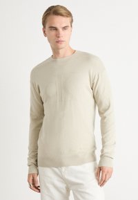 Maglione beige in tessuto morbido con colletto a giro e polsini a coste. Il davanti presenta un logo in rilievo discreto.