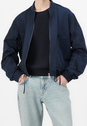 Person trägt eine marineblaue Bomberjacke über einem schwarzen Pullover, Hände in den Taschen hellblauer Jeans, zugeschnitten am Hals und an der Taille.