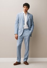 Young man wearing a light blue suit, white shirt, and brown loafers standing against a beige wall with one hand in his pocket.