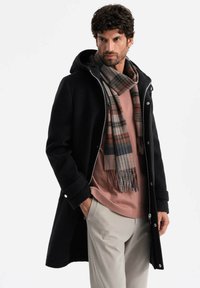 Abrigo negro con capucha, cierre frontal y botones a presión. Llevado con una camisa rosa y una bufanda de cuadros en tonos grises, marrones y azules. Pantalones de color claro.