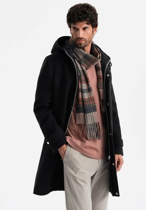 Abrigo negro con capucha, cierre frontal y botones a presión. Llevado con una camisa rosa y una bufanda de cuadros en tonos grises, marrones y azules. Pantalones de color claro.