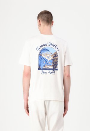 Osoba o krótkich włosach ubrana w biały t-shirt Tommy Hilfiger z niebiesko-beżowym tropikalnym nadrukiem i napisem New York na plecach.