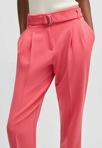 Růžové kalhoty na míru s širokým pásem a kovovou sponou, hladká textura a skladbový design, ukázané s rukama v kapsách.
