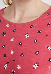 Camiseta de algodón en color coral con cuello redondo. Presenta estampados florales dispersos en azul marino y blanco. Textura suave, diseño casual.