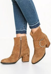 Botas de tobillo de ante marrón con tacón de bloque, cremallera lateral y un acento de cinturón con una hebilla metálica circular.