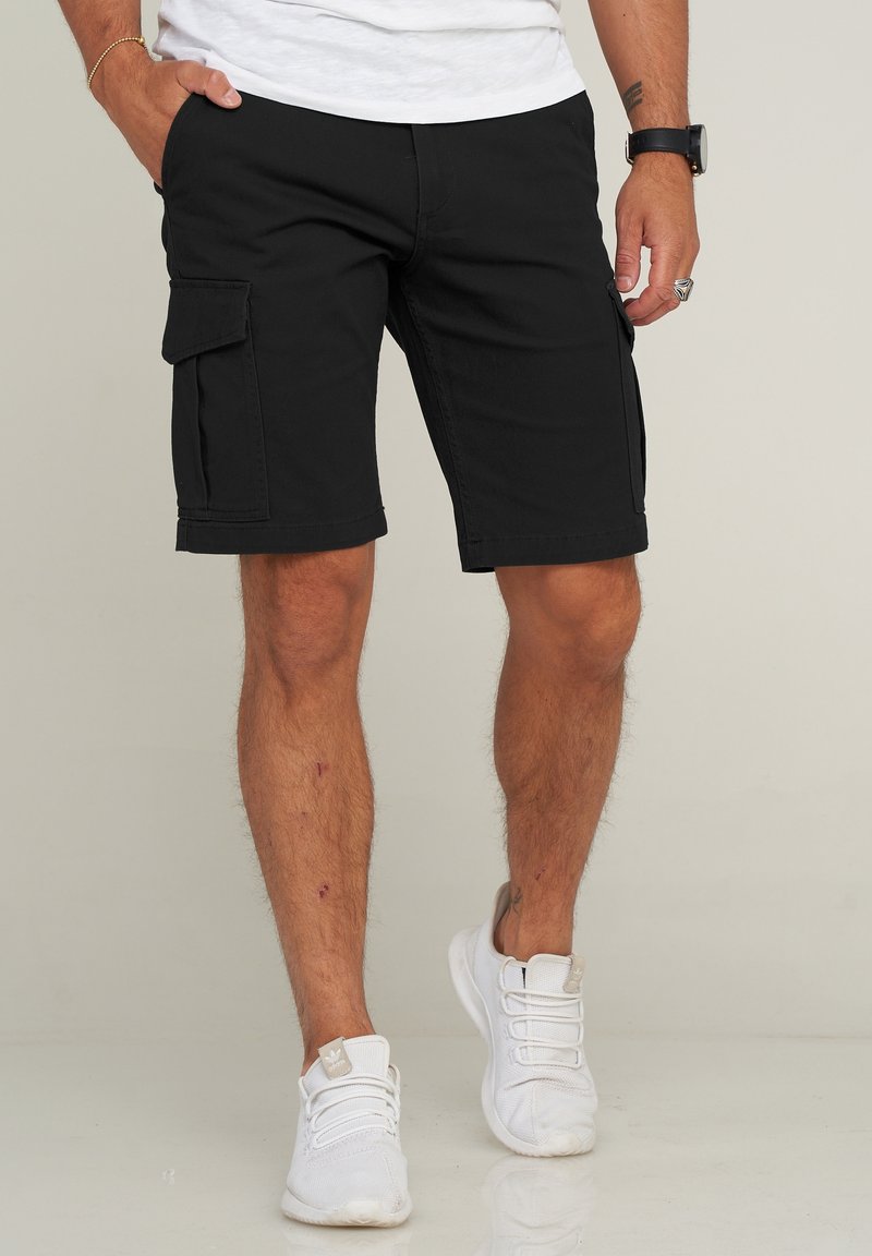 Shorts cargo noirs avec deux poches latérales, longueur genou, en tissu mélangé coton. Portés avec des baskets blanches sur une surface claire et réfléchissante.