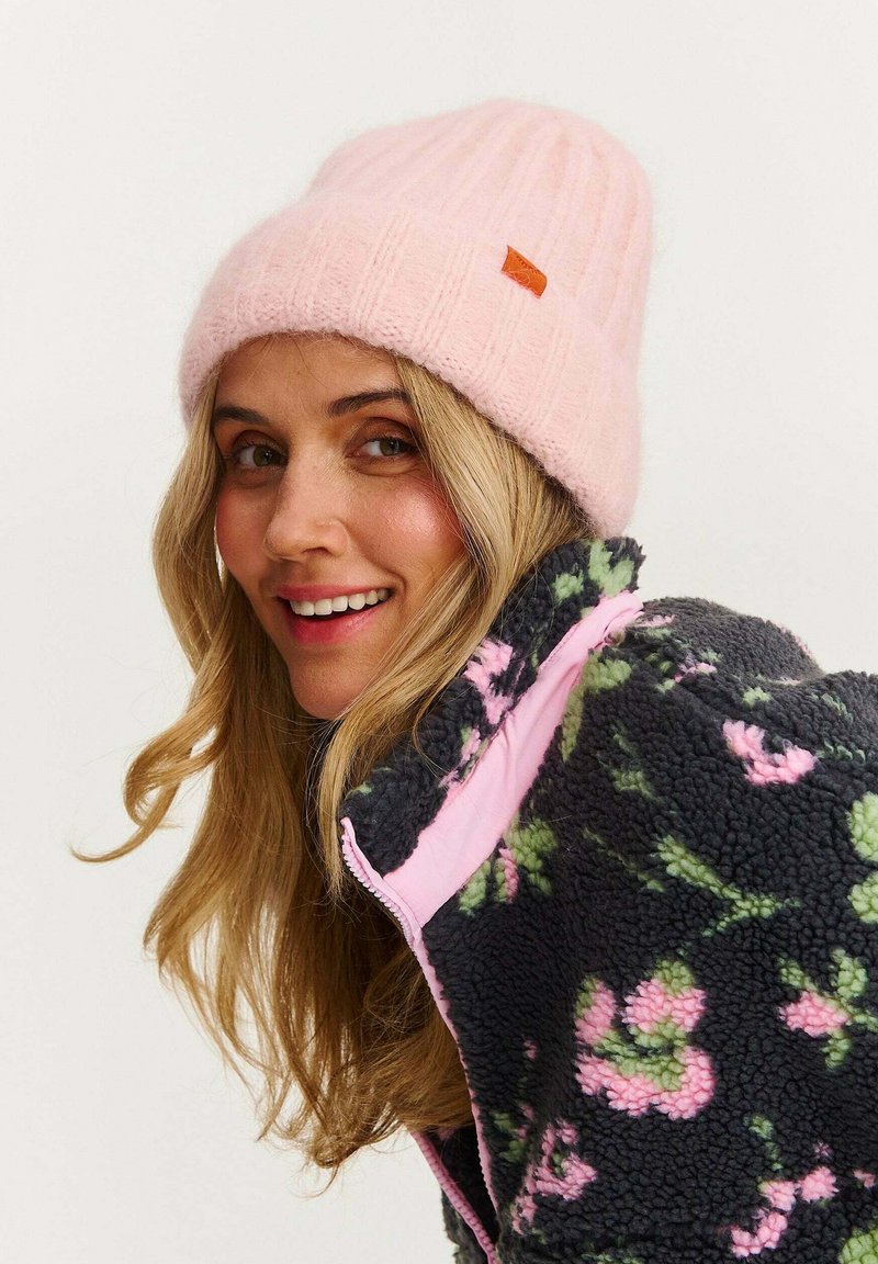 Svetloružová rebrovaná pletená čiapka s malou oranžovou nálepkou, nosená s tmavou mikinou s kvetinovým vzorom a ružovými a zelenými akcentmi.