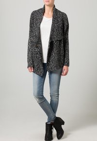 Cardigan à pois noir et blanc avec col châle, manches longues et deux poches avant, associé à un t-shirt blanc et un jean en denim.