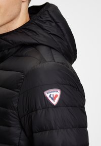 Svart puffjacka med huva, med kviltad textur och en framträdande Rossignol-logotyp på vänster axel.