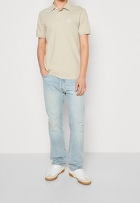 Homme portant un polo beige à manches courtes, un jean déchiré bleu clair et des baskets blanches, se tenant devant un fond clair uni.