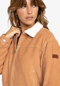 Kobaltcotton jack met warme bruine kleur en een fuzzy witte kraag, ritssluiting en een zijzak. Bevat een bruine leren logo patch.